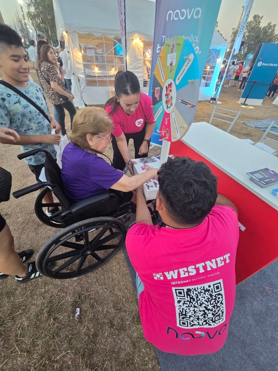 Integrante de Westnet ayudando a una vecina en silla de ruedas a girar la ruleta de premios del stand