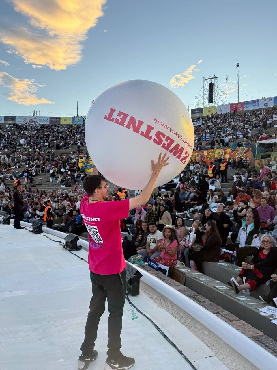 Integrante de Westnet sosteniendo una pelota inflable gigante de la marca frente a las tribunas del Frank Romero Day al atardecer